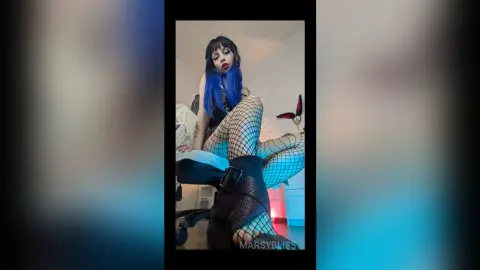 Woman with blue hair in fishnet tights and black outfit sitting on a bed in a moody bedroom setting.
