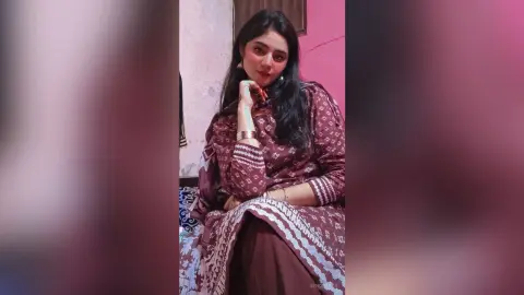 Young woman in a patterned maroon ethnic tunic sitting indoors with a thoughtful expression.