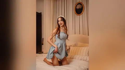 Elegant woman in a light blue summer dress posing on a bed in a cozy, sunlit bedroom.