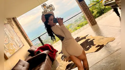 KendallRosy poses in a tight tan bodysuit while standing on a rug overlooking a scenic valley view.
