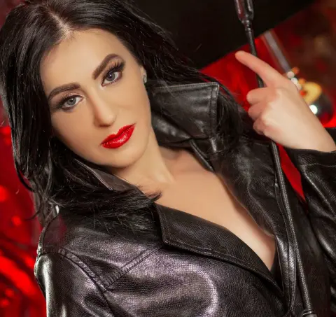 Glamorous woman in a black leather jacket posing with a microphone against a red background.