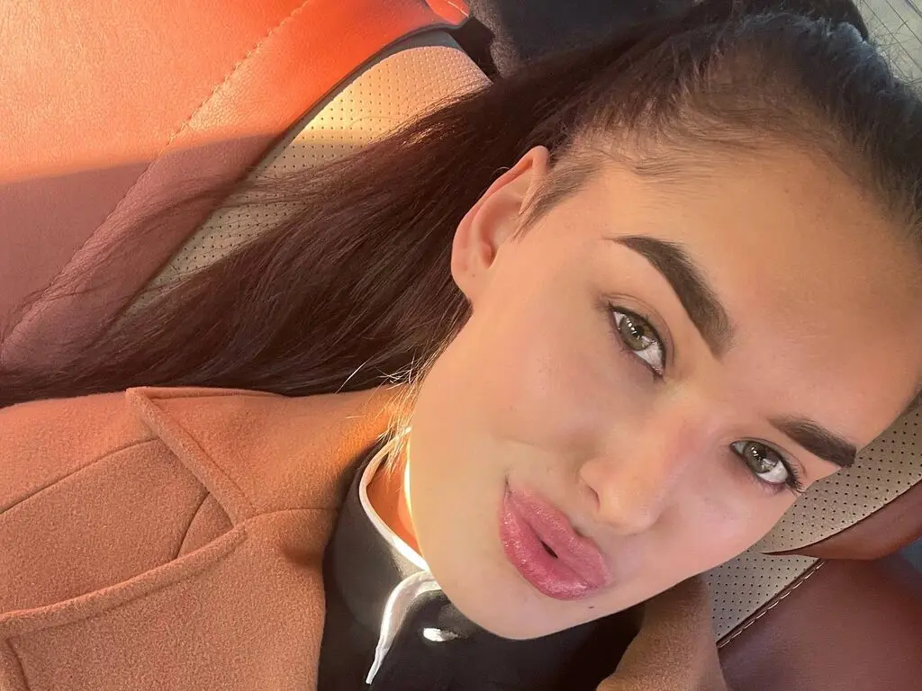EmelyMastorovo pouts her lips while wearing a brown coat and black shirt, posing in a car seat.