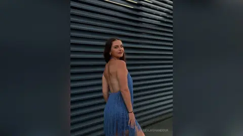 Elegant woman in a blue fringed backless dress posing against a dark slatted wall in a moody setting.