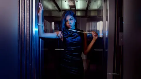 Elegant woman in a black satin dress posing in a moody, blue-lit elevator with a pearl necklace.