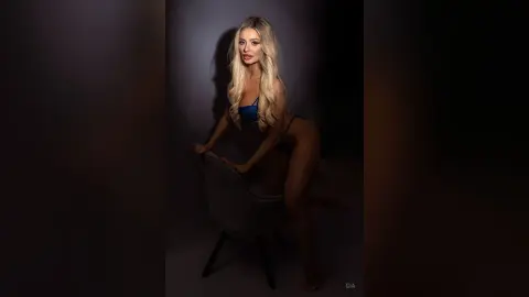 Gia poses naked behind a white chair, showing her long blonde hair and bare body against a dark background.
