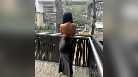 Woman in a backless black satin maxi dress standing on a city balcony overlooking urban buildings.