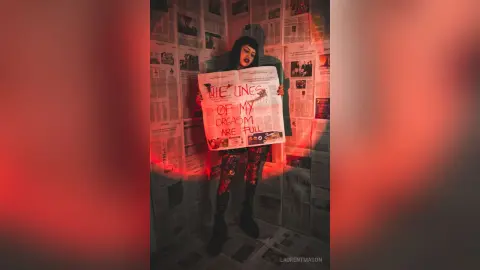 Moody portrait of a woman in dark clothing holding a newspaper sign in a room covered in newsprint.