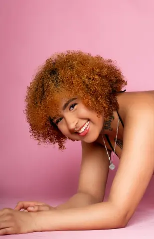 Smiling woman with curly ginger hair and nose ring posing against a pink background.