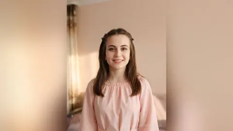 Smiling young woman in a pink blouse sitting in a bright, cozy room with soft natural lighting.