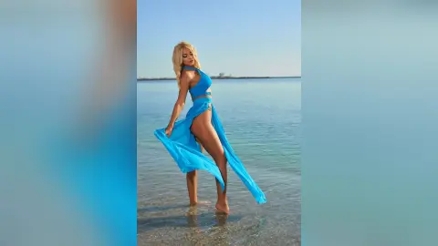 Anastasia stands in shallow ocean water wearing a long, bright blue slit dress that shows her bare legs.
