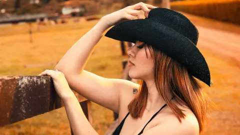 Young woman in a black sun hat relaxing by a rustic wooden fence during a warm golden hour sunset.