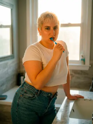 Young woman with short blonde hair in a white t-shirt and jeans brushing teeth in a bright, sunny bathroom.