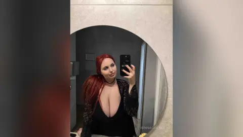 Redheaded woman in a black lace outfit taking a moody mirror selfie in a modern bathroom.