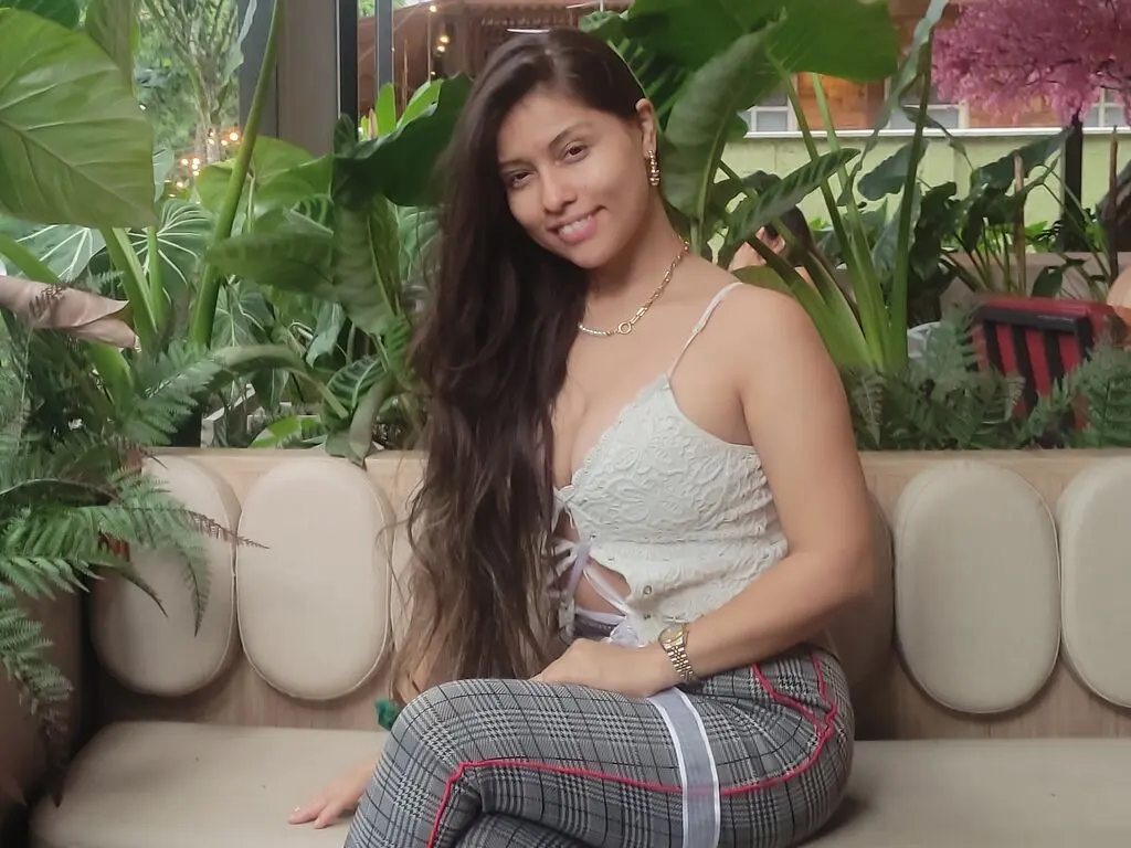 Smiling woman in a white lace camisole and grey plaid trousers sitting on a beige sofa in a lush garden setting.