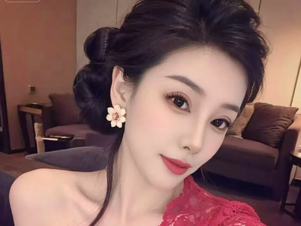 Elegant woman with dark hair in a bun wearing a red lace dress and flower earrings in a cozy living room.