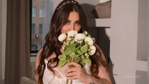 KeiraHarts poses with a bouquet of white flowers, wearing a white lace bra and a pearl headband in her long, wavy brown hair.