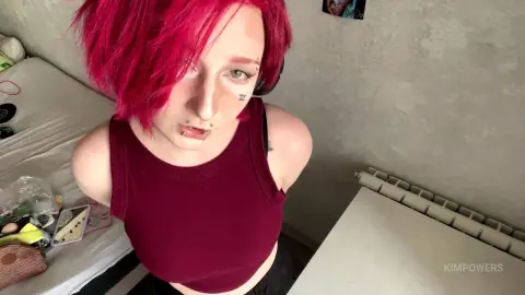 Woman with bright pink hair and facial piercings wearing a maroon tank top in a cozy bedroom setting.