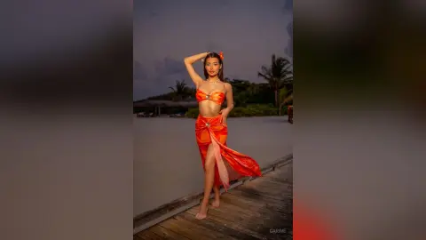 Elegant woman in an orange tropical outfit posing on a wooden pier at sunset by the ocean.