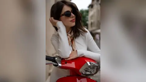 Jessie poses behind a red scooter wearing a white long-sleeved top and dark sunglasses.