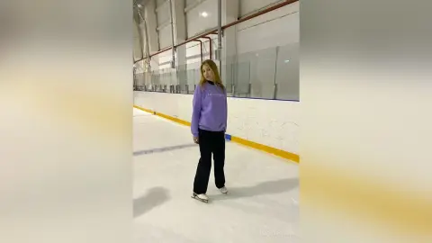 Young woman in a purple sweatshirt and black pants ice skating in an indoor arena.