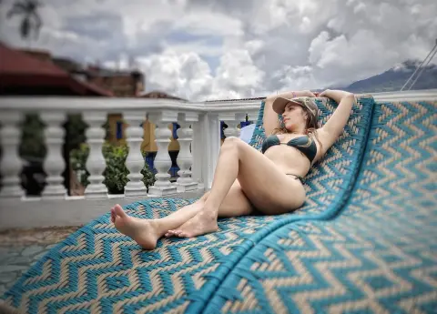 Relaxed woman in a bikini lounging on a patterned sunbed on a sunny balcony overlooking a scenic view.