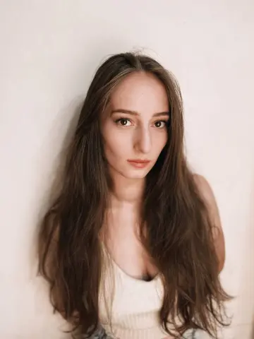 Young woman with long brown hair wearing a white ribbed tank top against a neutral background.