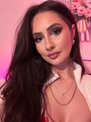 Glamorous woman with long dark hair and gold jewelry posing in soft pink lighting.