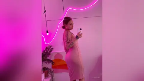 LucyLoy poses for a mirror selfie in a tight pink dress under bright pink neon lights.