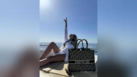 Blonde woman in a white outfit relaxing on a boat deck by the ocean with a designer Christian Dior tote bag.
