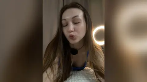 Young woman with long brown hair in a blue tank top making a kissy face in a cozy, dimly lit room.
