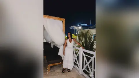 Elegant woman in a white maxi dress standing on a luxury balcony at night near tropical palm trees.