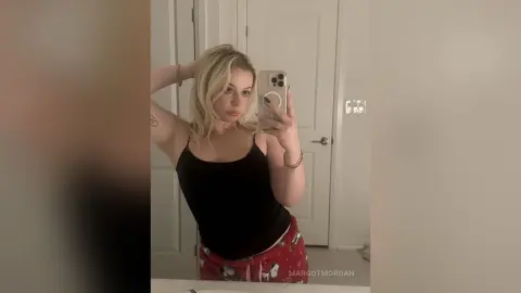 Blonde woman in a black tank top taking a casual mirror selfie in a bright bathroom.