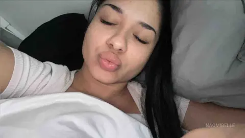 Young woman with long dark hair relaxing in bed wearing a white t-shirt in a cozy, peaceful mood.