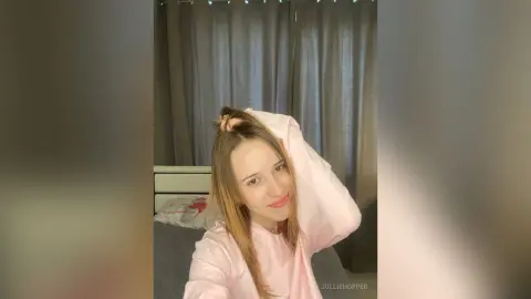 Smiling young woman in a pink long-sleeve shirt relaxing in a cozy bedroom setting.