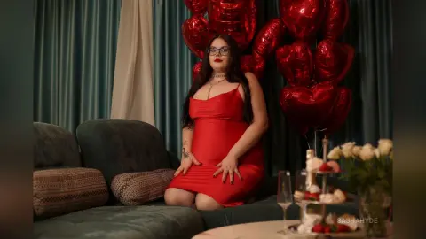 Pregnant woman in a red satin dress sitting on a green sofa surrounded by red heart balloons.