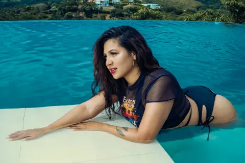 Curvy woman in a black graphic crop top relaxing by a bright blue swimming pool on a sunny day.
