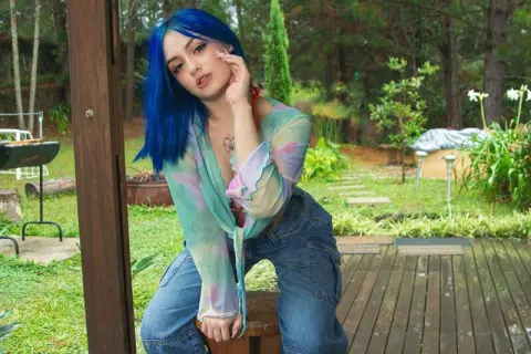 Blue-haired woman in a pastel tie-dye blouse sitting on a wooden deck in a lush green garden.