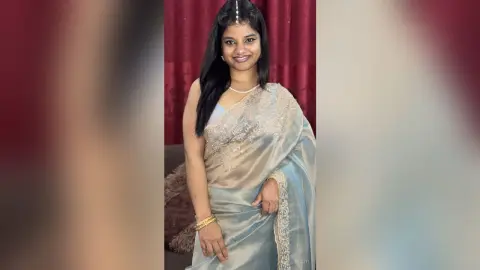 Sejuti smiles while wearing a silver silk saree and a pearl necklace, posing against a red curtain background.