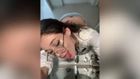 Young woman in a white shirt wearing glasses leaning over a bathroom sink with a playful expression.