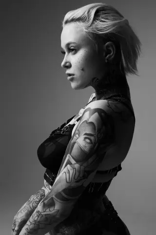 Black and white portrait of a blonde woman with extensive arm and neck tattoos wearing a black bodysuit.