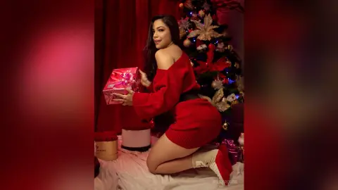Smiling woman in a red sweater holding a pink gift box near a decorated Christmas tree in a festive setting.