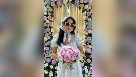 Stylish woman in a white floral sundress and headscarf holding a pink peony bouquet by a floral arch.