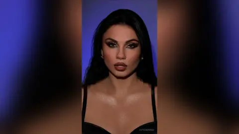 CutemEmma poses with long dark hair and heavy eye makeup, wearing a black bra against a blue background.