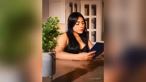 Young woman in a black top reading a book at a wooden table near a potted plant in a cozy indoor setting.