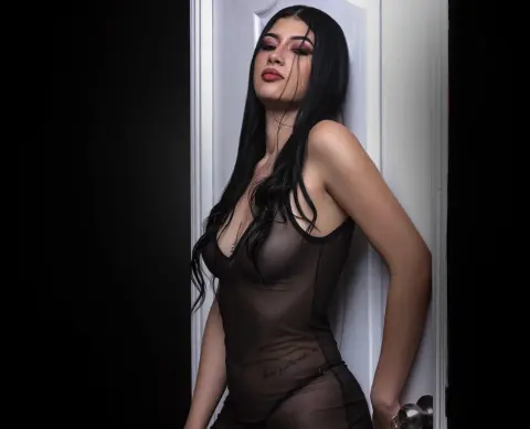 Elegant woman in a sheer black bodysuit posing against a white door in moody, dramatic lighting.