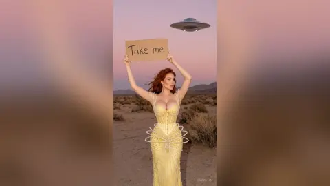 Redheaded woman in a glamorous yellow corset gown holding a 'Take me' sign in a desert landscape at sunset.