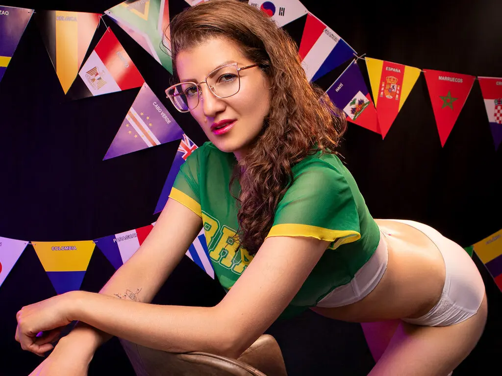 AnnaJhonson wears glasses and a green jersey, posing while leaning forward to show her white panties and bare skin against a background of colorful flags.