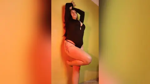 EsmeraldMoon poses against a yellow wall wearing a black long-sleeve bodysuit, lifting one leg up while holding her hands behind her head.