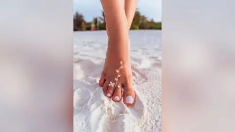 Elie shows off her feet with white nail polish while standing on a sandy beach.