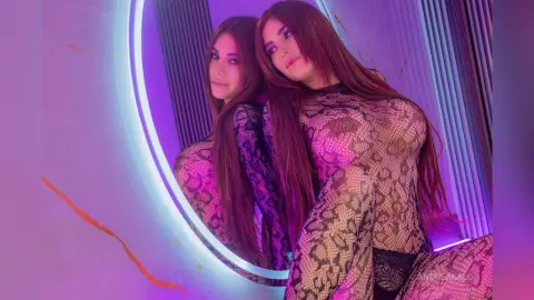 AndreaKalo poses in a sheer, black snakeskin-patterned bodysuit, showing her long brown hair and cleavage in a purple-lit room with a circular mirror.